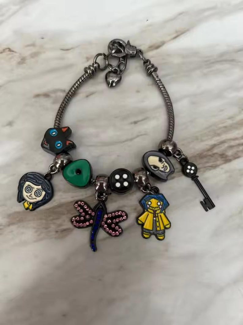 May include: Silver charm bracelet featuring various character charms. Charms include a black cat, a green button, a dragonfly, a key, and character faces. The bracelet has a lobster clasp closure.