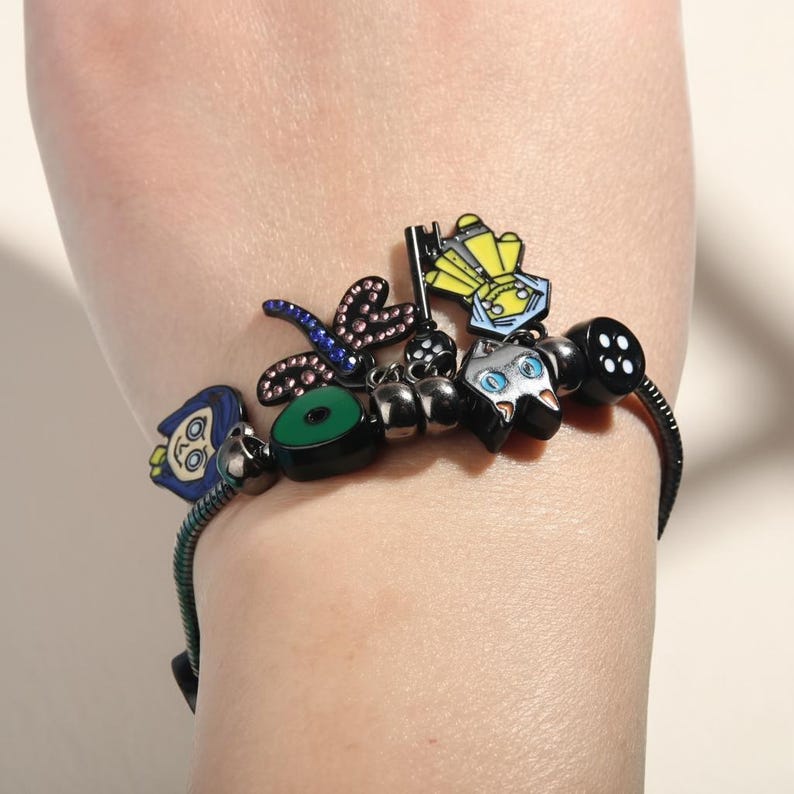 May include: A black charm bracelet with various colorful charms. Charms include a dragonfly, a cat, a key, and a green circle. The bracelet is worn on a wrist.