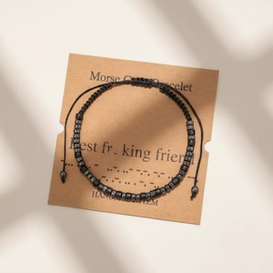 May include: A black Morse code bracelet with the message "Best f*cking friend" on a brown card. The bracelet is made of small, dark beads strung on a black cord. The card also reads "Morse Code Bracelet" and "Handmade Item."