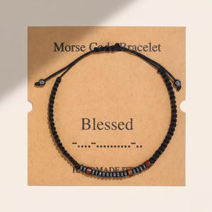 May include: A black Morse code bracelet with the word "Blessed" printed on a brown card. The bracelet features a braided cord and small beads in shades of brown and gray.