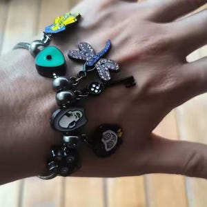 May include: A silver charm bracelet with various decorative charms. Charms include a dragonfly with rhinestone accents, a key, a face, and other unique designs. The bracelet is on a person's wrist.