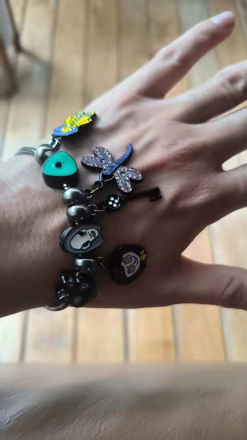 May include: A silver charm bracelet with various decorative charms. Charms include a dragonfly with rhinestone accents, a key, a face, and other unique designs. The bracelet is on a person's wrist.
