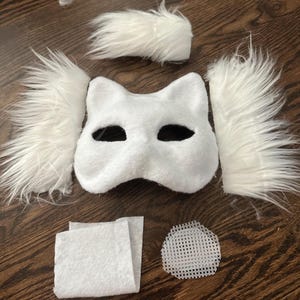 May include: White felt cat mask with black eye holes and white fur trim. The mask is laid out on a wooden surface with additional pieces of white felt and white fur.