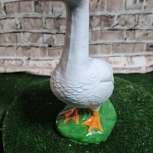 May include: A white goose figurine with an orange beak and feet, perched on a green base. The goose has a long neck and is painted with detailed feathers. The background is a brick wall.