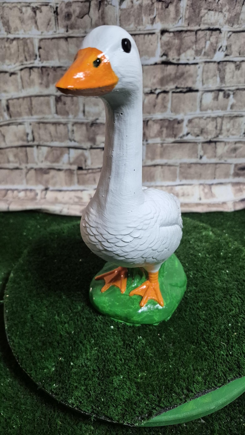 May include: A white goose figurine with an orange beak and feet, perched on a green base. The goose has a long neck and is painted with detailed feathers. The background is a brick wall.