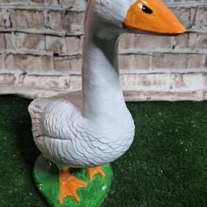 May include: A ceramic goose figurine, painted white with an orange beak and feet. The goose stands on a green base, set against a brick wall background. The figurine is a decorative item.