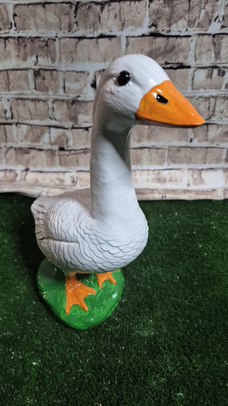 May include: A ceramic goose figurine, painted white with an orange beak and feet. The goose stands on a green base, set against a brick wall background. The figurine is a decorative item.