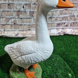 May include: A white goose statue with an orange beak and feet, perched on a green base. The goose has black eyes and detailed feather textures. The background features a brick wall and green turf.