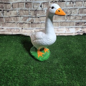 May include: A white goose figurine with an orange beak and feet stands on a green base. The goose has detailed feather textures and a long neck. The background features a brick-like pattern.