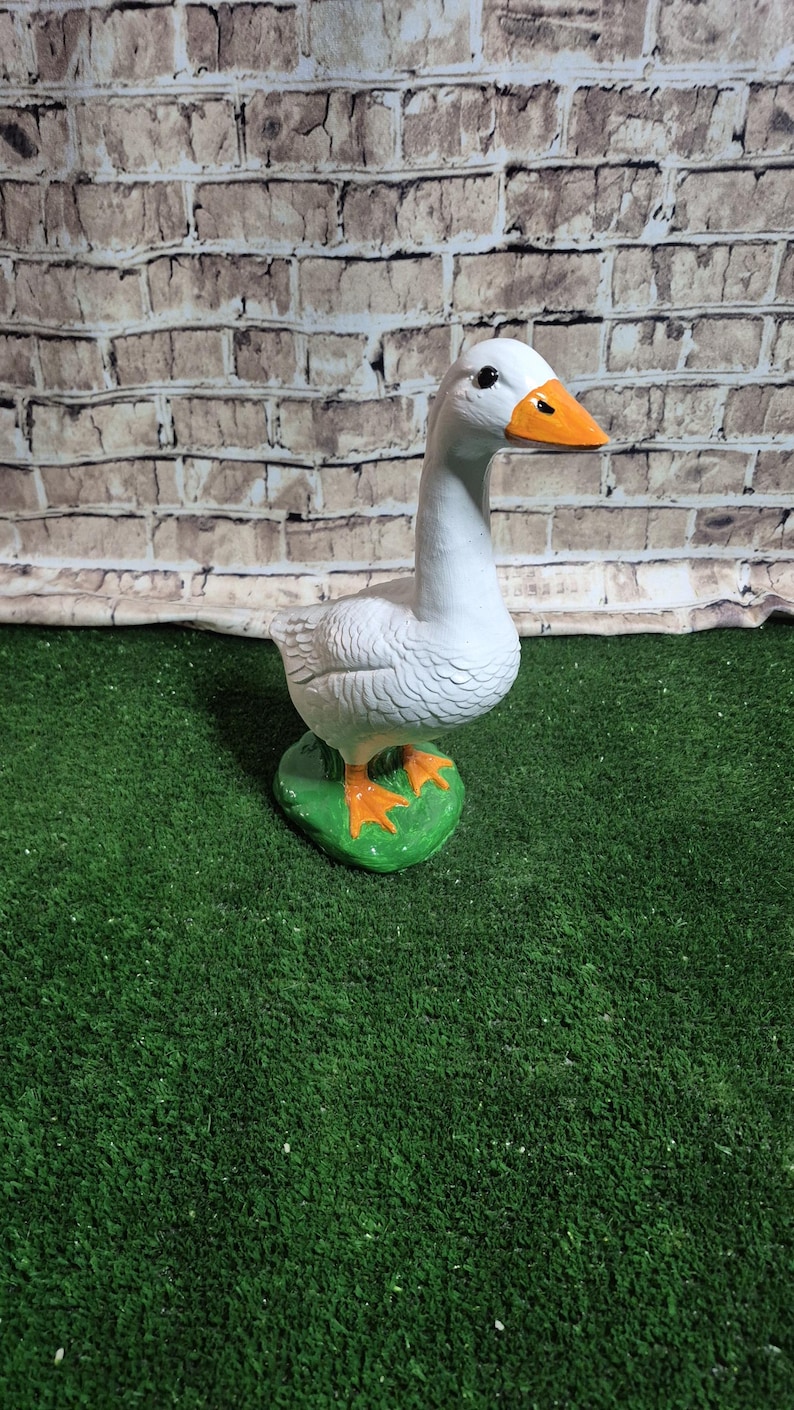 May include: A white goose figurine with an orange beak and feet stands on a green base. The goose has detailed feather textures and a long neck. The background features a brick-like pattern.