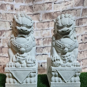 May include: Two matching, light gray Foo Dog statues. Each statue is seated on a decorative base with intricate carvings. The Foo Dogs have detailed features, including manes, collars, and paws. The background is a brick wall and green grass.