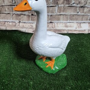 May include: A white goose figurine with an orange beak and feet, standing on a green base. The goose has black eyes and is painted with a textured finish. The background is a brick wall.