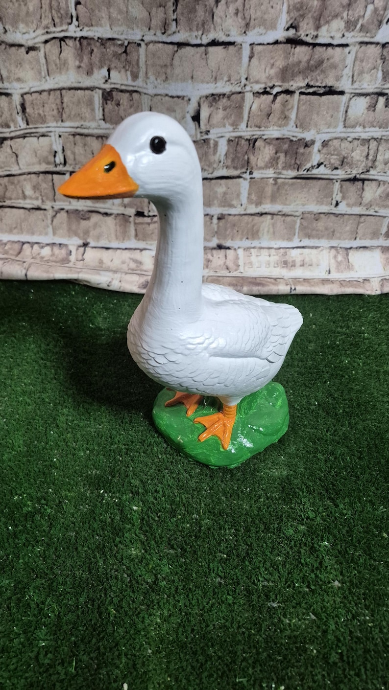 May include: A white goose figurine with an orange beak and feet, standing on a green base. The goose has black eyes and is painted with a textured finish. The background is a brick wall.