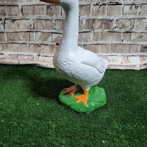 May include: A white goose statue with an orange beak and feet, standing on a green base. The goose has a long neck and is painted with a glossy finish. The background is a brick wall.