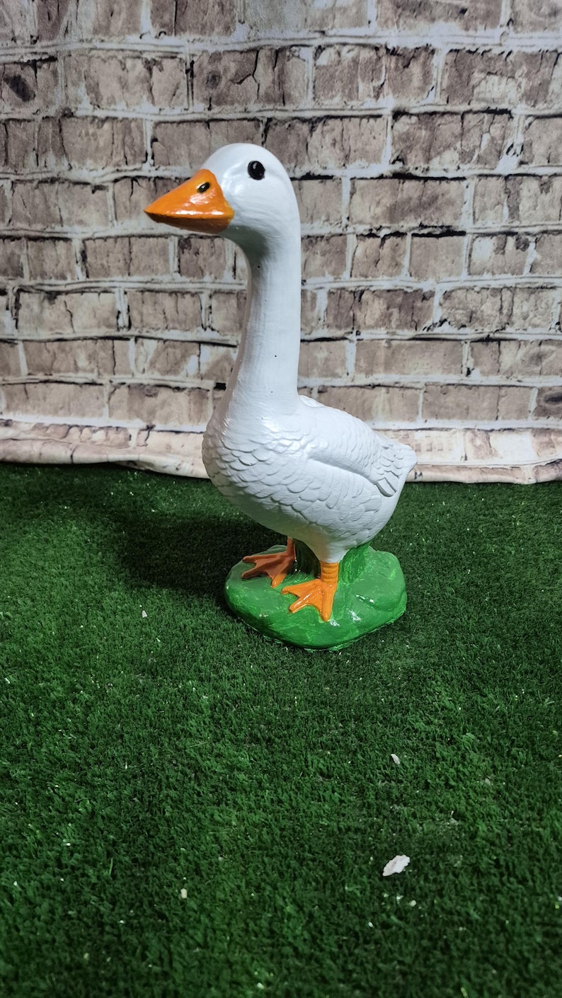 May include: A white goose statue with an orange beak and feet, standing on a green base. The goose has a long neck and is painted with a glossy finish. The background is a brick wall.