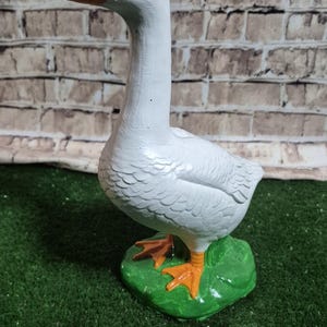 May include: A white goose figurine with an orange beak and feet, standing on a green base. The goose has a long neck and is set against a brick wall background. The figurine is a decorative item.