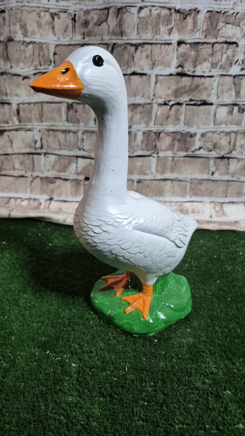 May include: A white goose figurine with an orange beak and feet, standing on a green base. The goose has a long neck and is set against a brick wall background. The figurine is a decorative item.