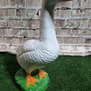 May include: A white goose figurine with an orange beak and feet, standing on a green base. The goose has black eyes and is set against a brick wall background. The figurine is a decorative item.