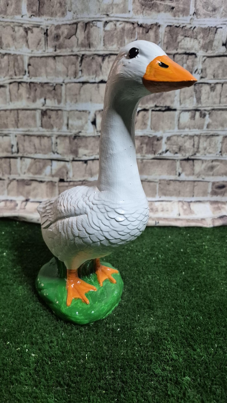 May include: A white goose figurine with an orange beak and feet, standing on a green base. The goose has black eyes and is set against a brick wall background. The figurine is a decorative item.