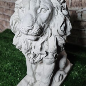 May include: A detailed, gray concrete lion statue. The lion is seated on a rectangular base, with a detailed mane and facial features. The statue is set against a brick wall and green artificial grass.