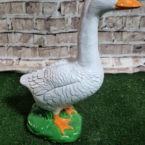 May include: A white goose figurine with an orange beak and feet, standing on a green base. The goose has detailed feather textures and black eyes. The background features a brick-patterned wall.