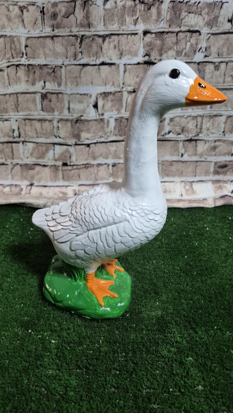 May include: A white goose figurine with an orange beak and feet, standing on a green base. The goose has detailed feather textures and black eyes. The background features a brick-patterned wall.