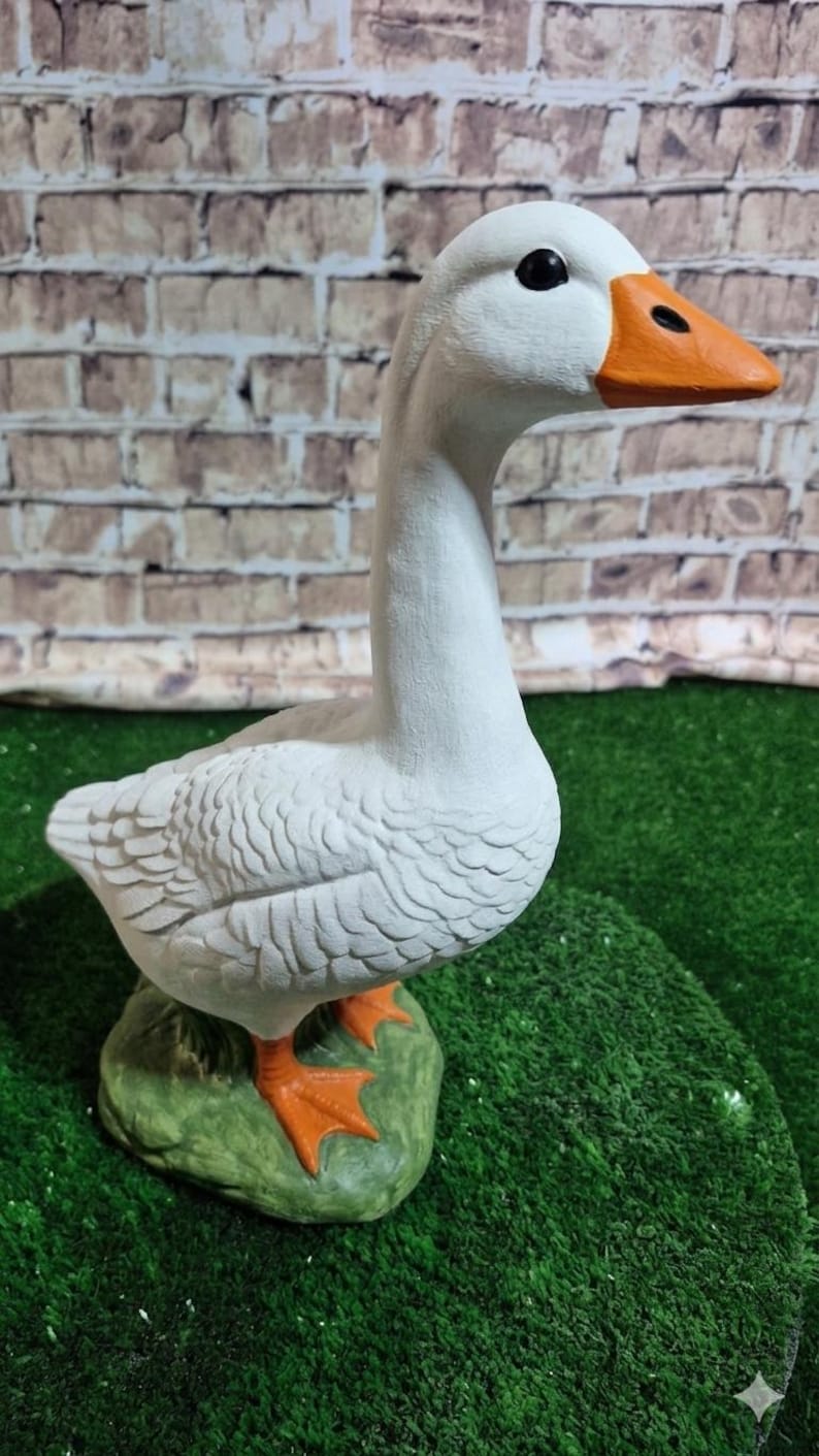 May include: A white goose statue with an orange beak and feet, perched on a green base. The goose has black eyes and detailed feather textures. The background features a brick wall and green turf.