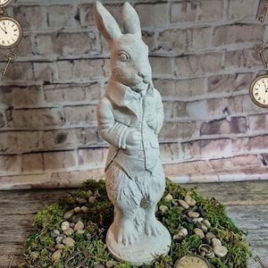 May include: A concrete statue of a rabbit wearing a jacket and vest, standing on a bed of moss and small stones. A vintage pocket watch and key are also present. The background is a brick wall.