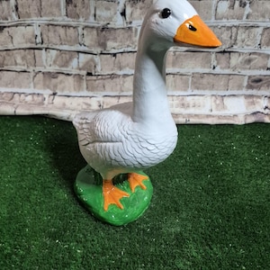 May include: A white goose statue with an orange beak and feet, standing on a green base. The goose has detailed feather textures and is set against a brick wall background.