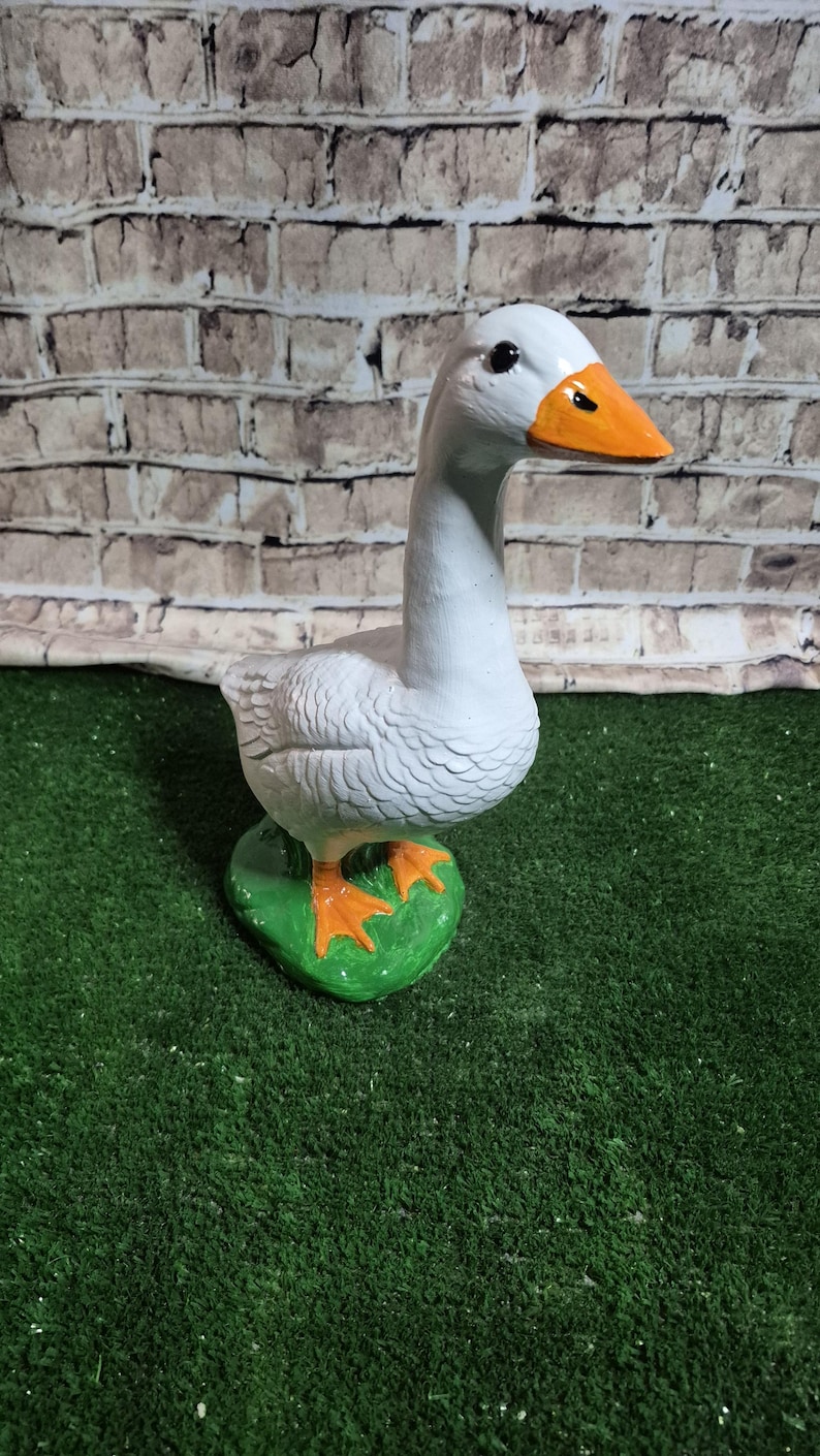 May include: A white goose statue with an orange beak and feet, standing on a green base. The goose has detailed feather textures and is set against a brick wall background.