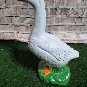 May include: A white goose figurine with an orange beak and feet, and black eyes. The goose stands on a green base, set against a brick wall background. The figurine is a decorative item.