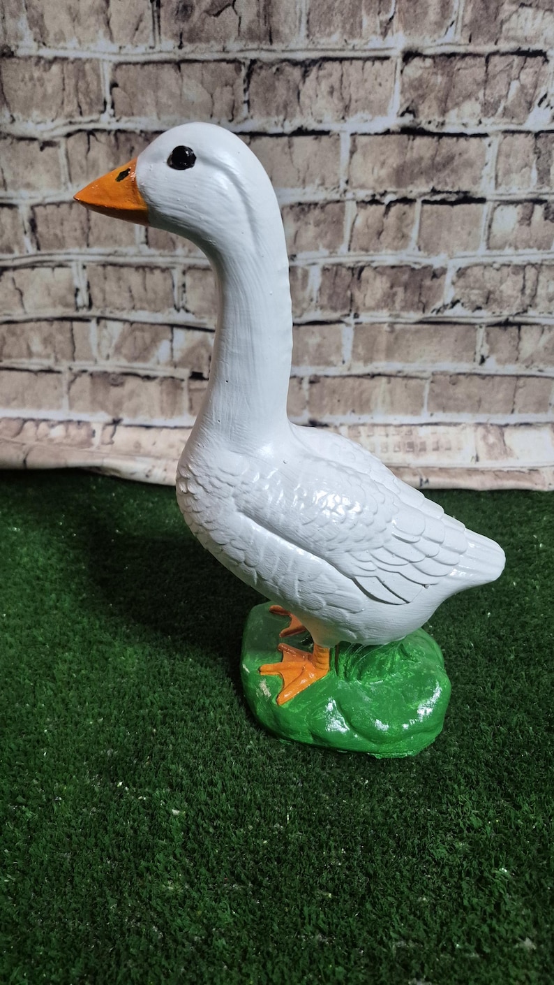 May include: A white goose figurine with an orange beak and feet, and black eyes. The goose stands on a green base, set against a brick wall background. The figurine is a decorative item.
