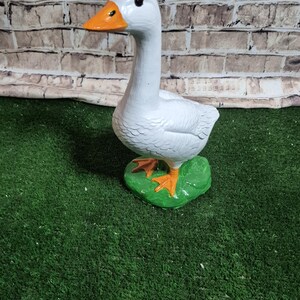 May include: A white goose figurine with an orange beak and feet, standing on a green base. The goose has black eyes and is set against a brick wall background. The figurine is a decorative item.