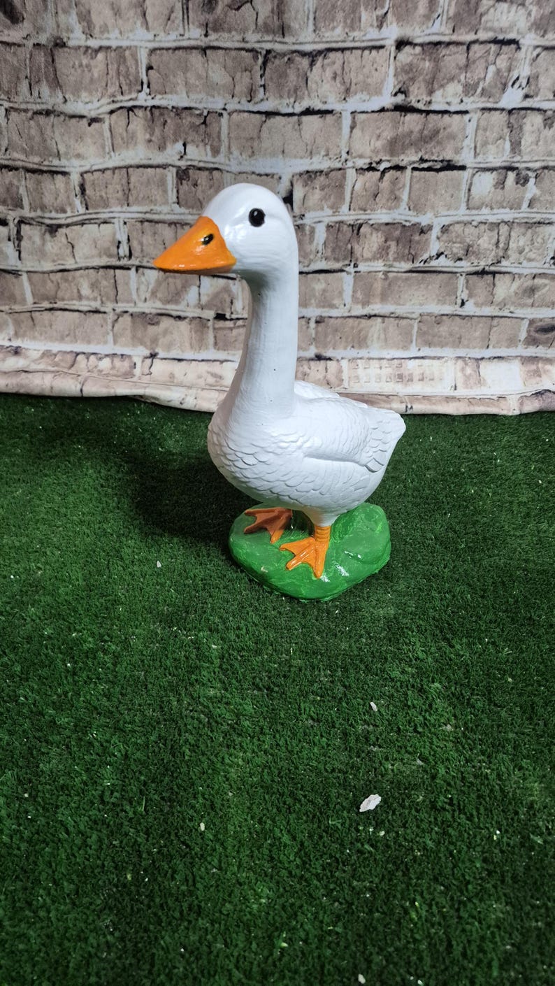 May include: A white goose figurine with an orange beak and feet, standing on a green base. The goose has black eyes and is set against a brick wall background. The figurine is a decorative item.
