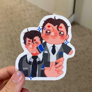 May include: A sticker featuring a cartoon illustration of two characters. The character on the left is wearing a blue and gray suit with a black tie and the text "RK800". The character on the right is wearing a white shirt, a black tie, and a gray suit. The character on the right is holding the character on the left's hand.