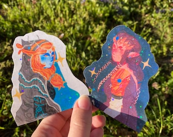 Könnte beinhalten: Zwei holografische Aufkleber mit Fantasy-Kunst. Einer zeigt einen blauhäutigen Charakter mit orangefarbenem Haar und einem schwarzen Umhang. Der andere zeigt einen Charakter mit rotem Haar und einem Sternenhintergrund. Beide Aufkleber haben ein glitzerndes Finish.