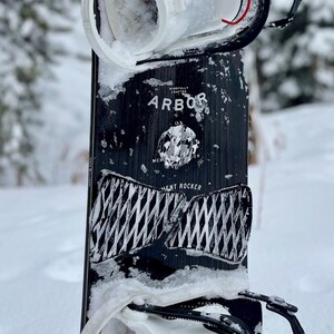 May include: A black snowboard with white text that reads "Mindfully Crafted Arbor" and a white graphic pattern. The snowboard has a white binding and is covered in snow.