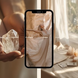 Może przedstawiać: Smartfon wyświetlający zdjęcie kobiety w zwiewnej białej sukni stojącej w pustynnym krajobrazie. Telefon leży na stole z świecą, kryształami i notesem.