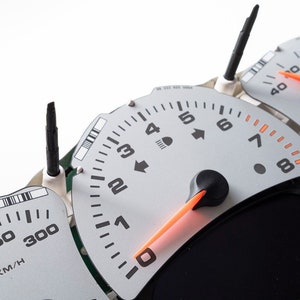May include: Close-up of a car's dashboard with three gauges. The speedometer reads up to 300 km/h, the tachometer reads up to 8, and the third gauge is unreadable. The gauges are silver with black markings and orange needles.