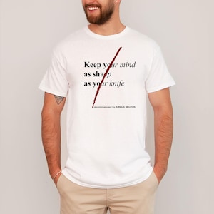 May include: White t-shirt with a red slash through the text "Keep your mind as sharp as your knife." The text "recommended by JUNIUS BRUTUS" is below the slash.