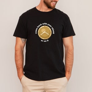 Könnte beinhalten: Ein schwarzes T-Shirt mit einem goldenen Schild-Motiv und dem Text "Komm mit deinem Schild zurück oder darauf."