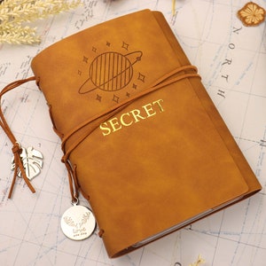 Könnte beinhalten: Ein braunes Lederjournal mit einem goldenen Prägemuster eines Planeten mit Sternen und dem Wort "SECRET". Das Journal hat einen Lederriemenverschluss und einen silbernen Anhänger mit einem Blattdesign.