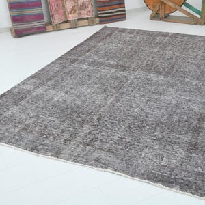 May include: A large, rectangular, distressed-look rug in shades of gray and brown. The rug has a faded, vintage appearance with a detailed pattern. The edges are finished with a lighter border. The rug is placed on a white floor.