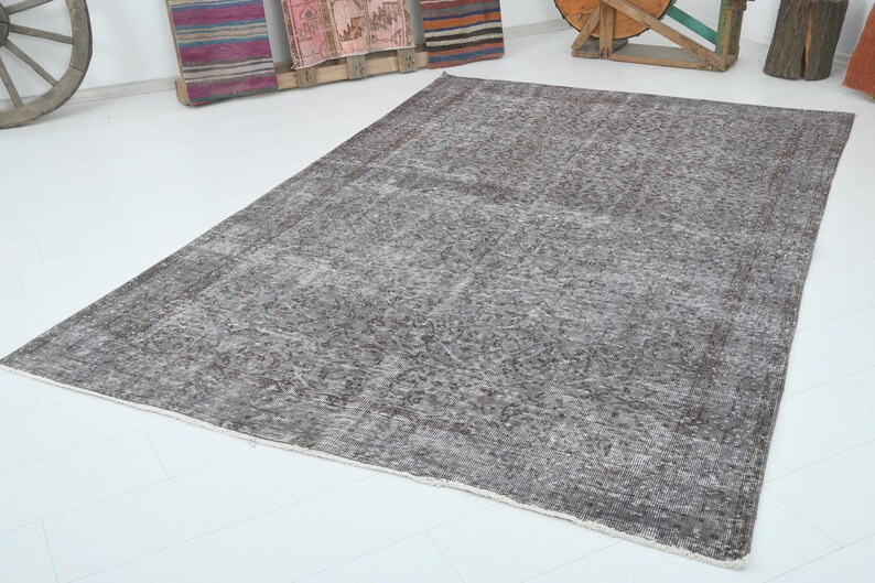 May include: A large, rectangular, distressed-look rug in shades of gray and brown. The rug has a faded, vintage appearance with a detailed pattern. The edges are finished with a lighter border. The rug is placed on a white floor.
