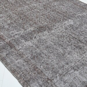 May include: A distressed, rectangular rug with a faded, vintage appearance. The rug features a gray and brown color palette with intricate floral patterns. The edges have a darker border, and the rug is placed on a white surface.