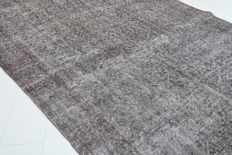 May include: A distressed, rectangular rug with a faded, vintage appearance. The rug features a gray and brown color palette with intricate floral patterns. The edges have a darker border, and the rug is placed on a white surface.
