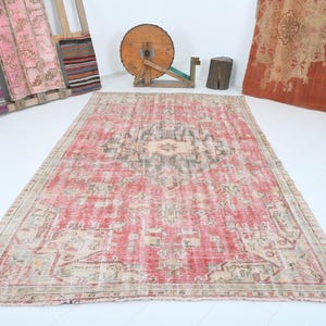May include: A rectangular, distressed red rug with a central medallion design in beige and gray. The rug has a faded, vintage appearance with a beige border. Other textiles and wooden objects are in the background.