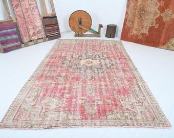 May include: A rectangular, distressed red rug with a central medallion design in beige and gray. The rug has a faded, vintage appearance with a beige border. Other textiles and wooden objects are in the background.