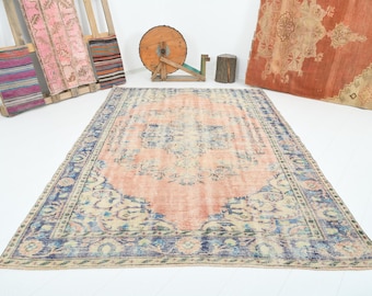 Pode incluir: Um tapete vintage grande e retangular com um campo central cor de pêssego e uma borda azul detalhada. O tapete apresenta um design desgastado e desbotado, sugerindo idade e uso. Outros têxteis e objetos de madeira estão no fundo.