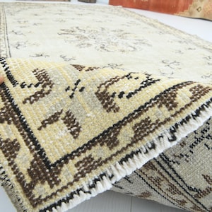 May include: Cream-colored vintage rug with a detailed floral pattern and a brown border. The rug's corner is folded, revealing a close-up of the intricate design and fringe. The rug is made of a soft, woven material.